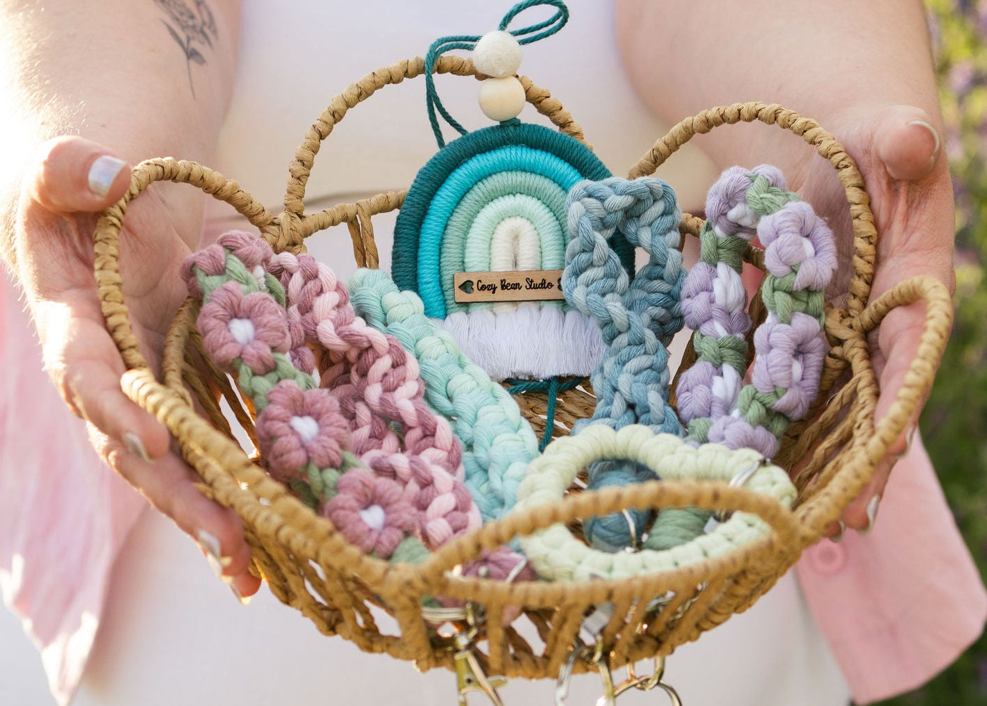 Heart-shaped woven basket with colorful floral and spiral key wristlet designs and teal rainbow diffuser held by a person in a blurred natural setting.
